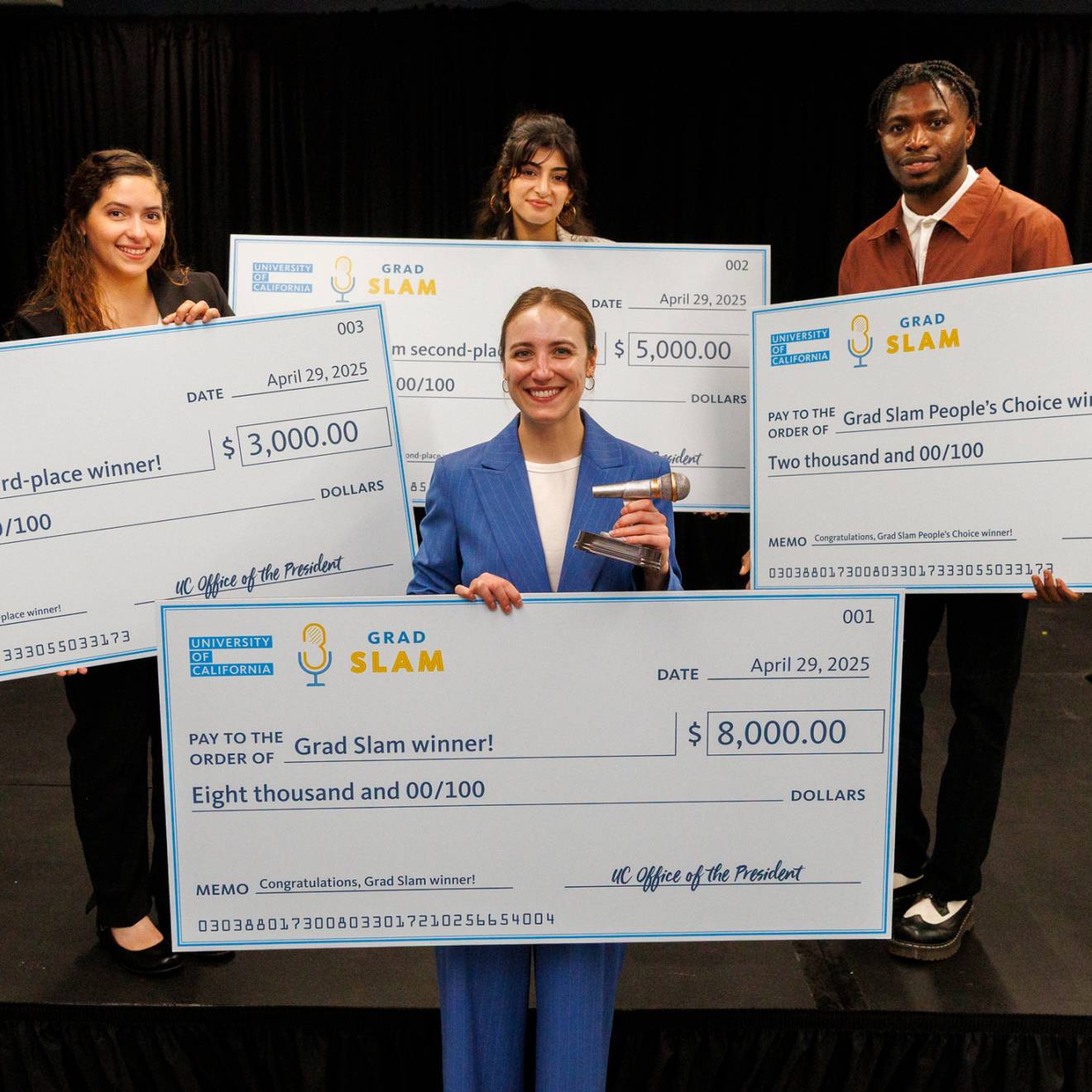 Four people, one in the foreground, three in the background, hold up giant checks
