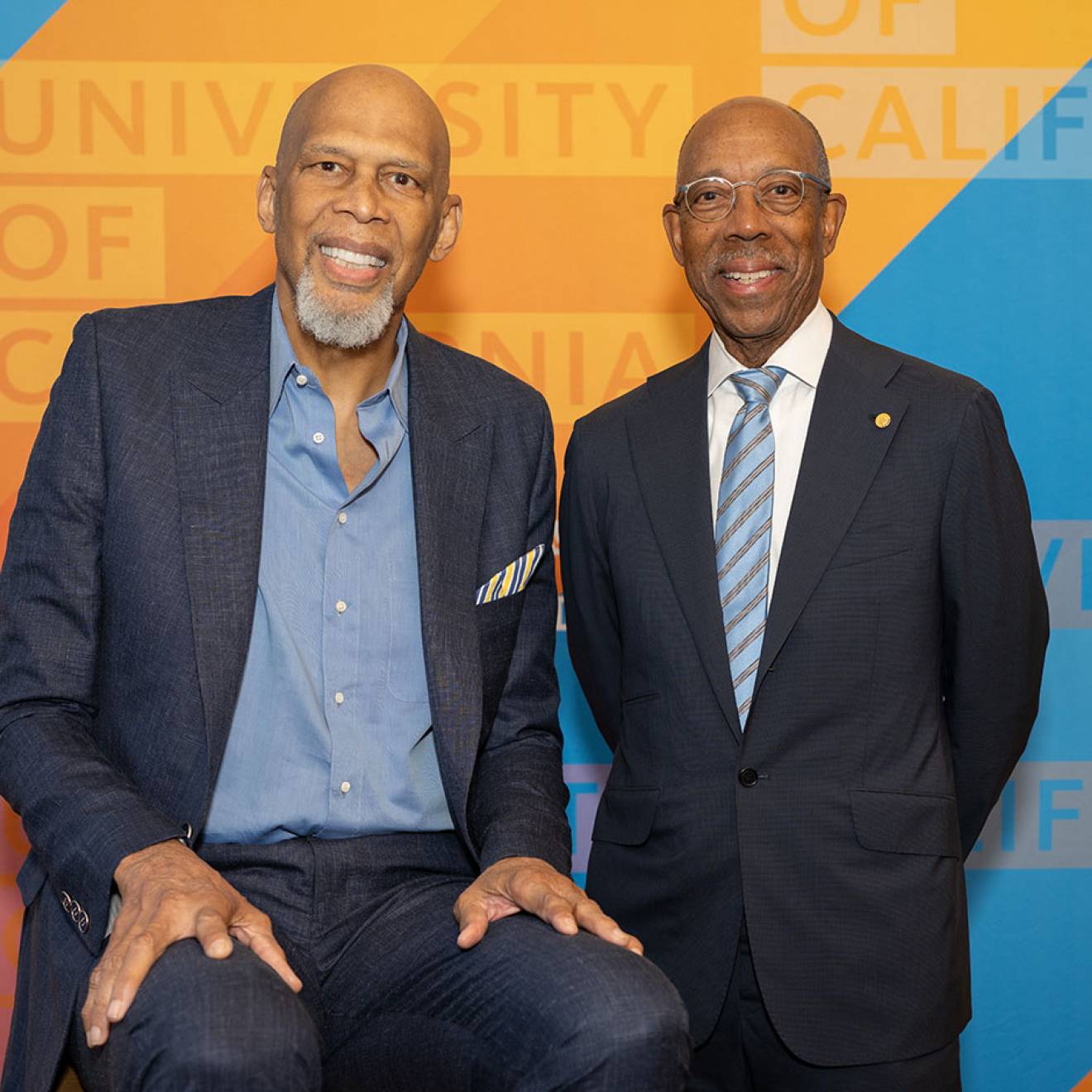 Kareem Abdul-Jabbar and UC President Michael Drake, M.D., smile for a portrait, standing in front of a University of California-branded backdrop