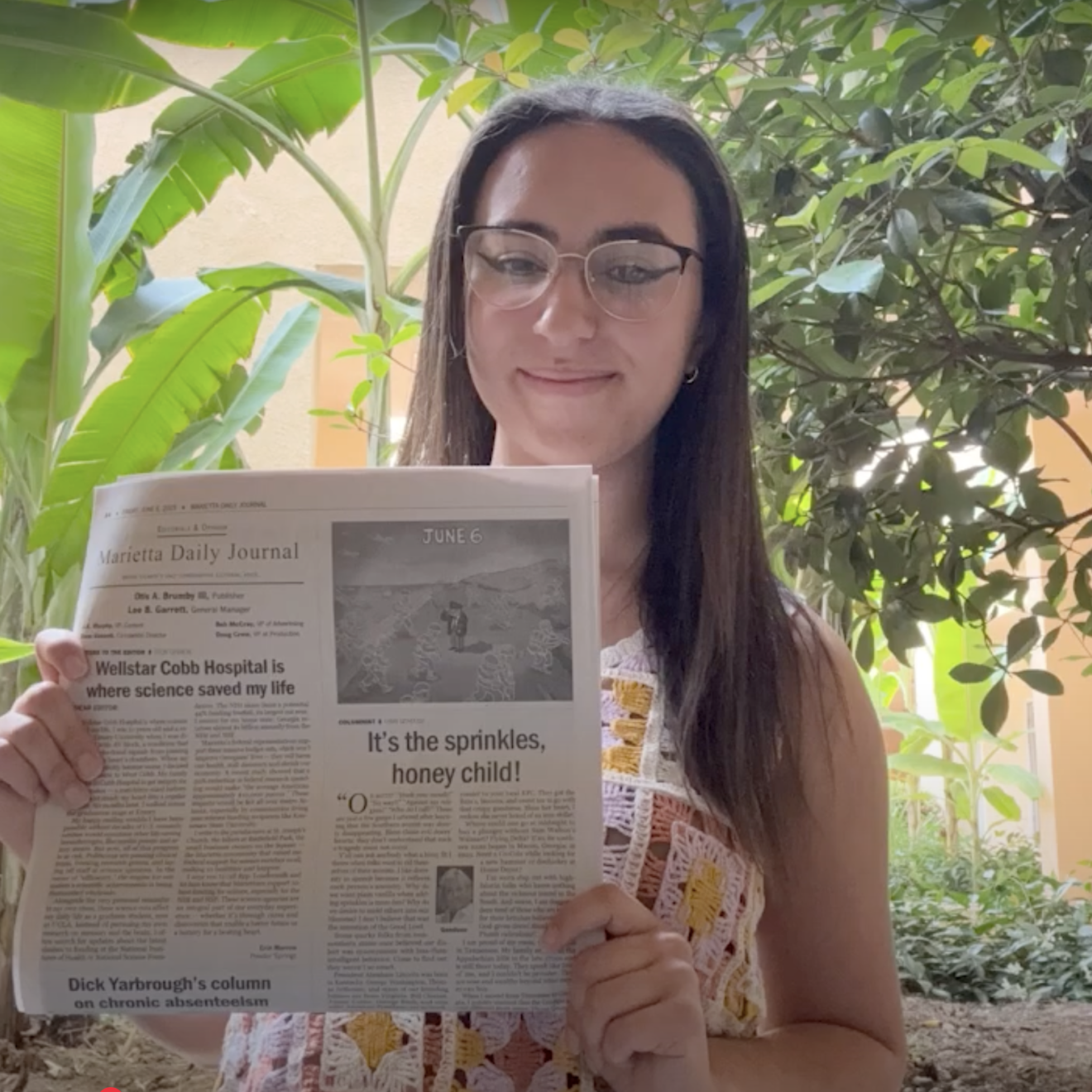 A person with long hair and glasses poses holding a newspaper