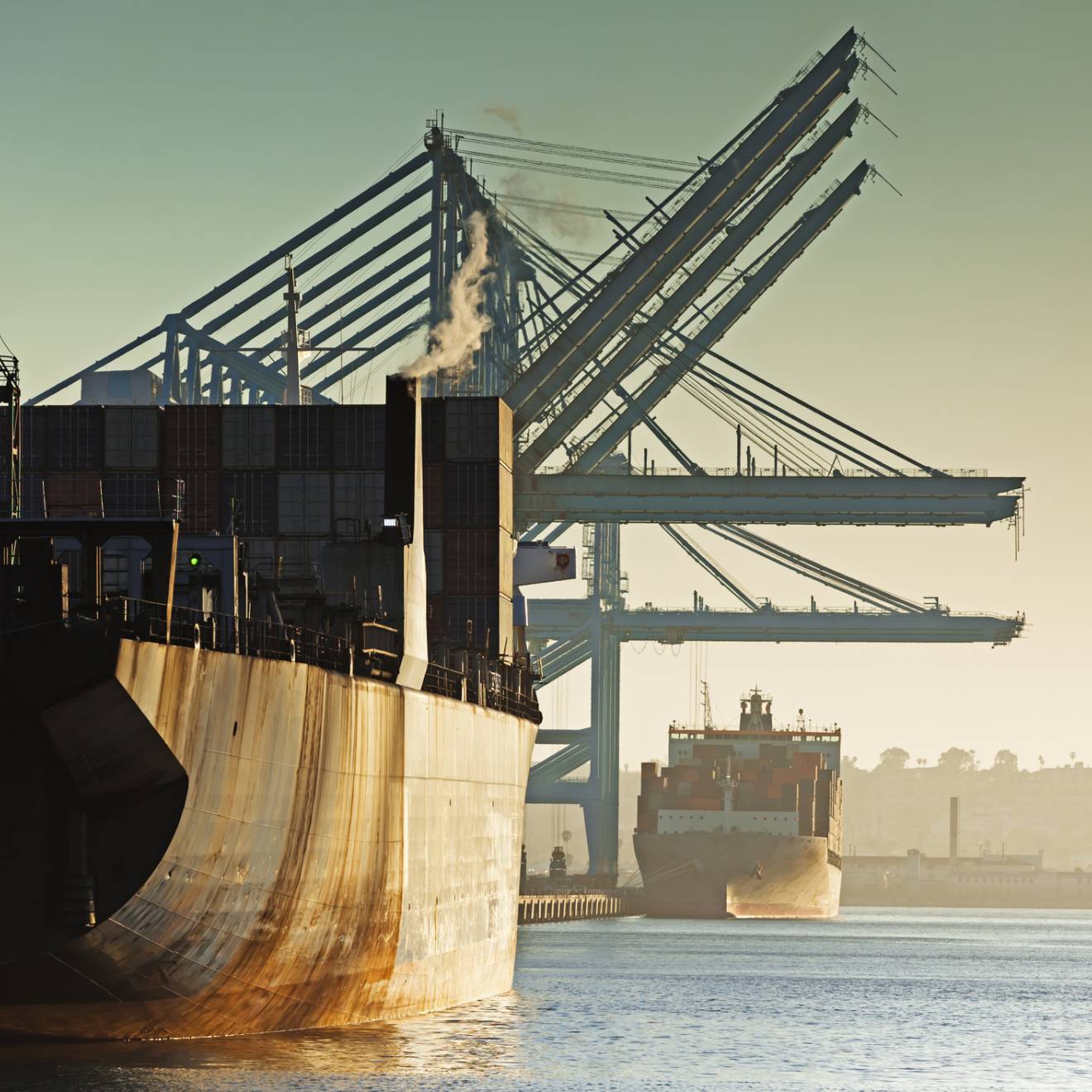 Two rusty container ships in a row, one behind, being loaded at a port during the evening