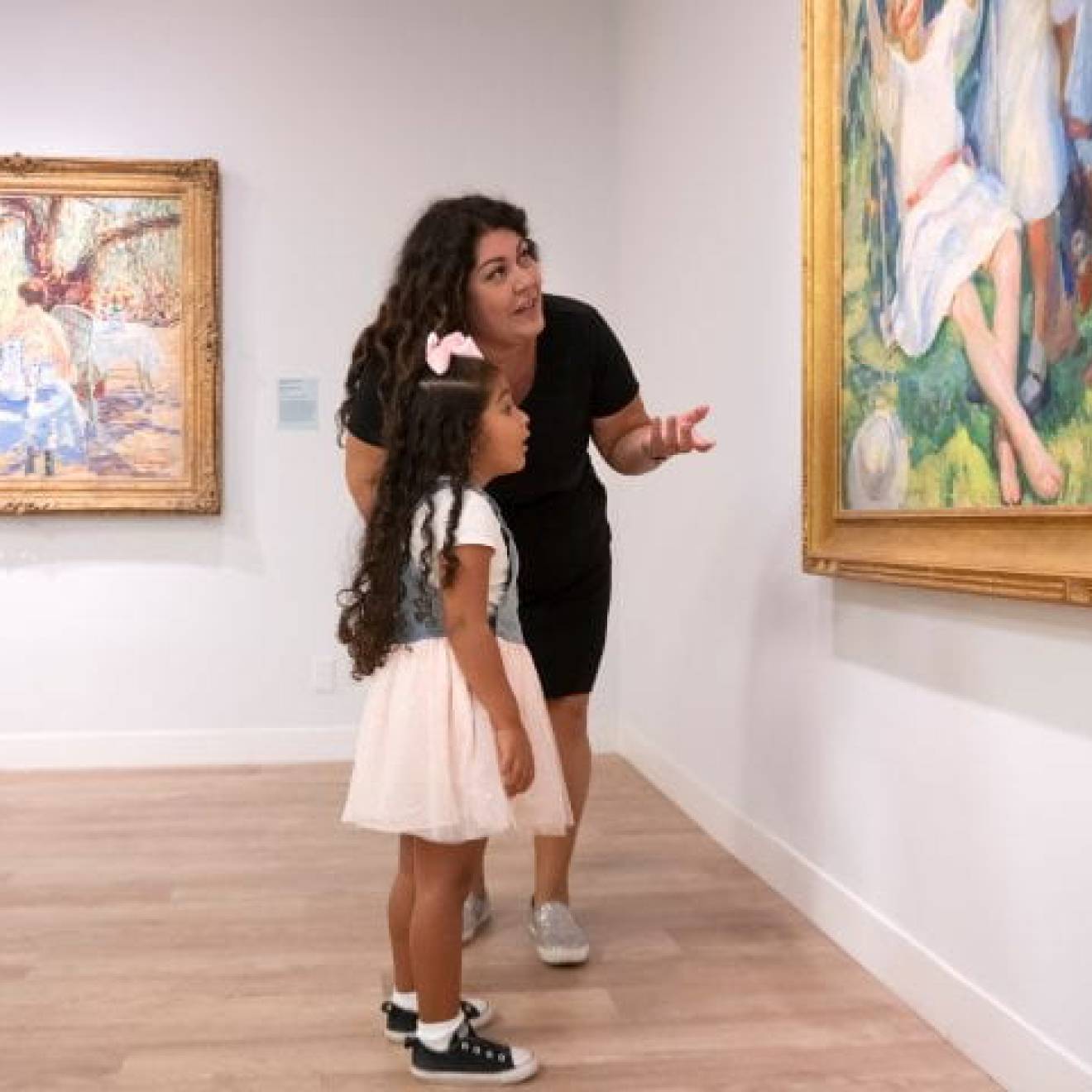 A mother and her young daughter talking about a painting they are looking in a museum gallery.