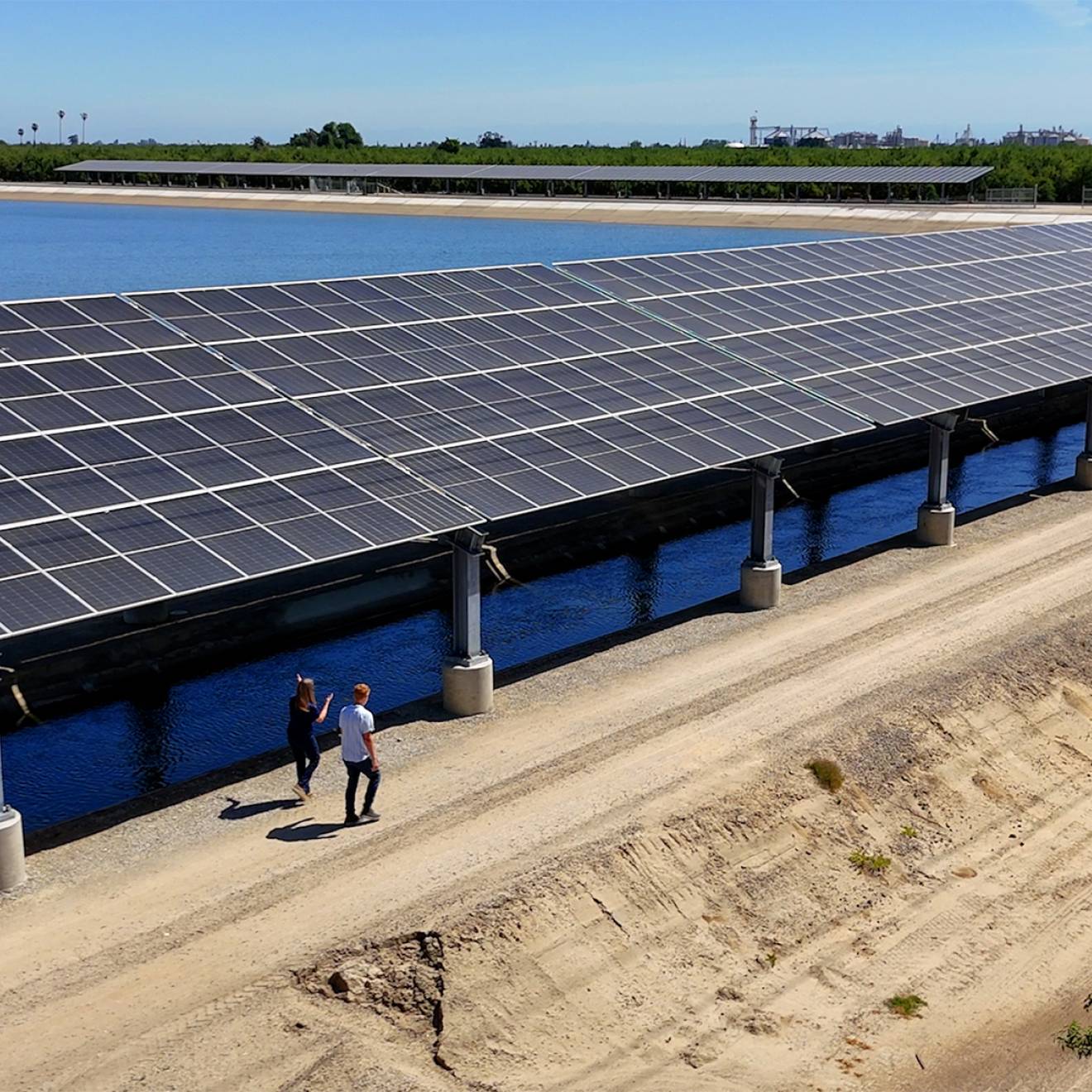 Two people walk next to a long set of solar panels over a canal