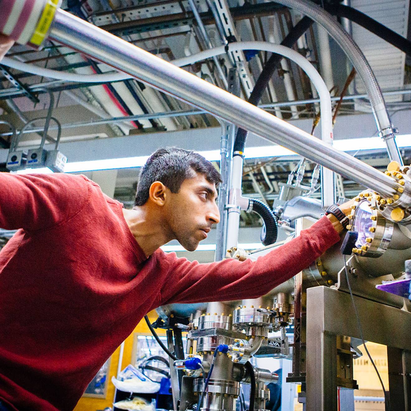 A grad student focuses intently as he slides a three-foot-long steel bar into a large apparatus made of tubes and wires, in a lab full of similar such apparatuses. 