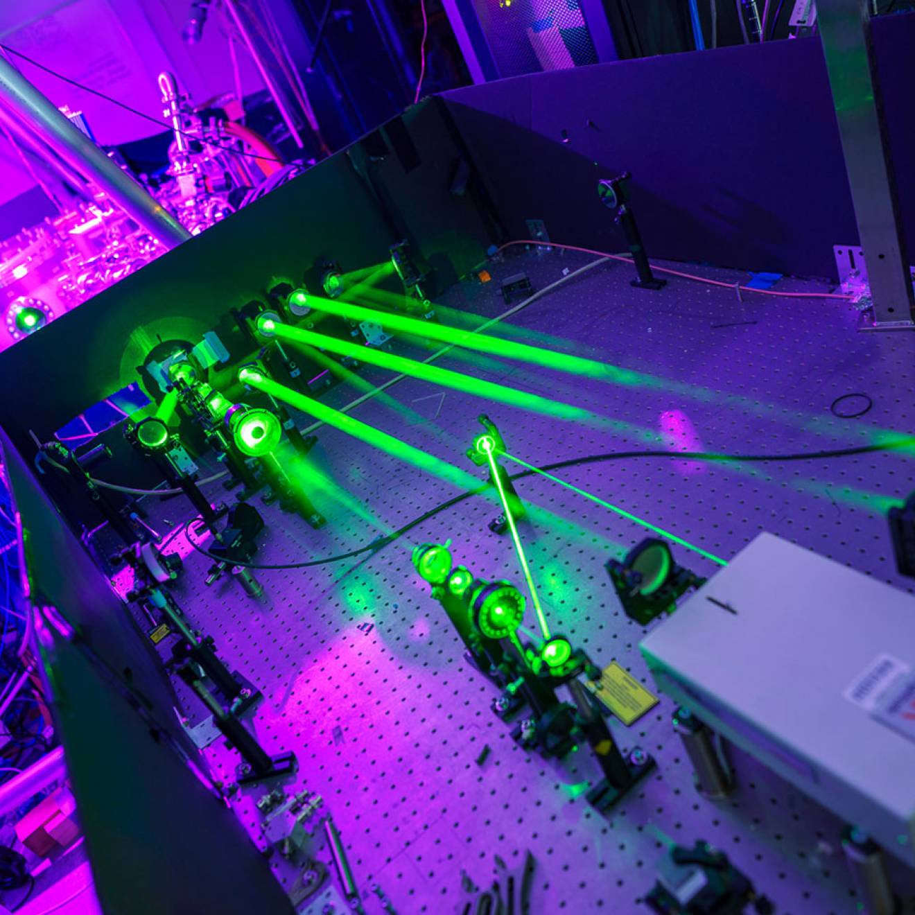 A laboratory setup with multiple bright green laser beams projected across an optical table lined with mirrors and instruments, illuminated by purple and blue ambient light, showing advanced experimental physics equipment.