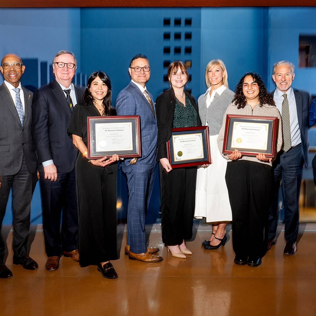 About ten people pose for a photo wearing business attire, three holding plaques.