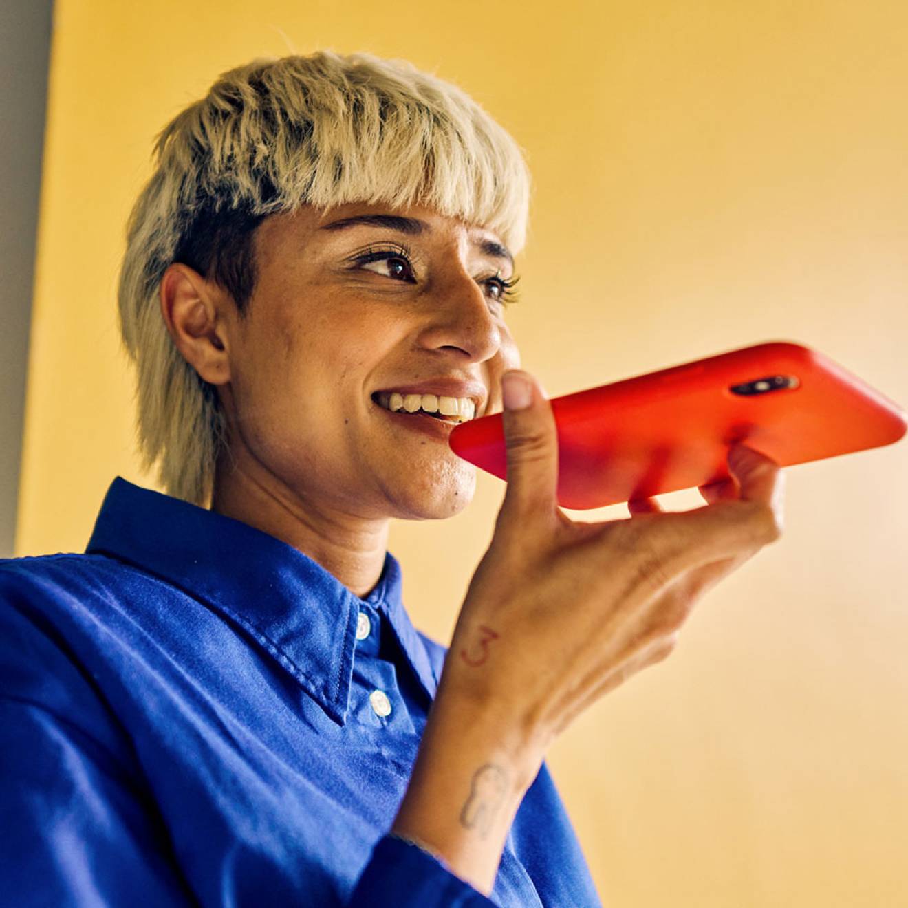 A young woman with a distinctive haircut speaks into a smart phone, smiling