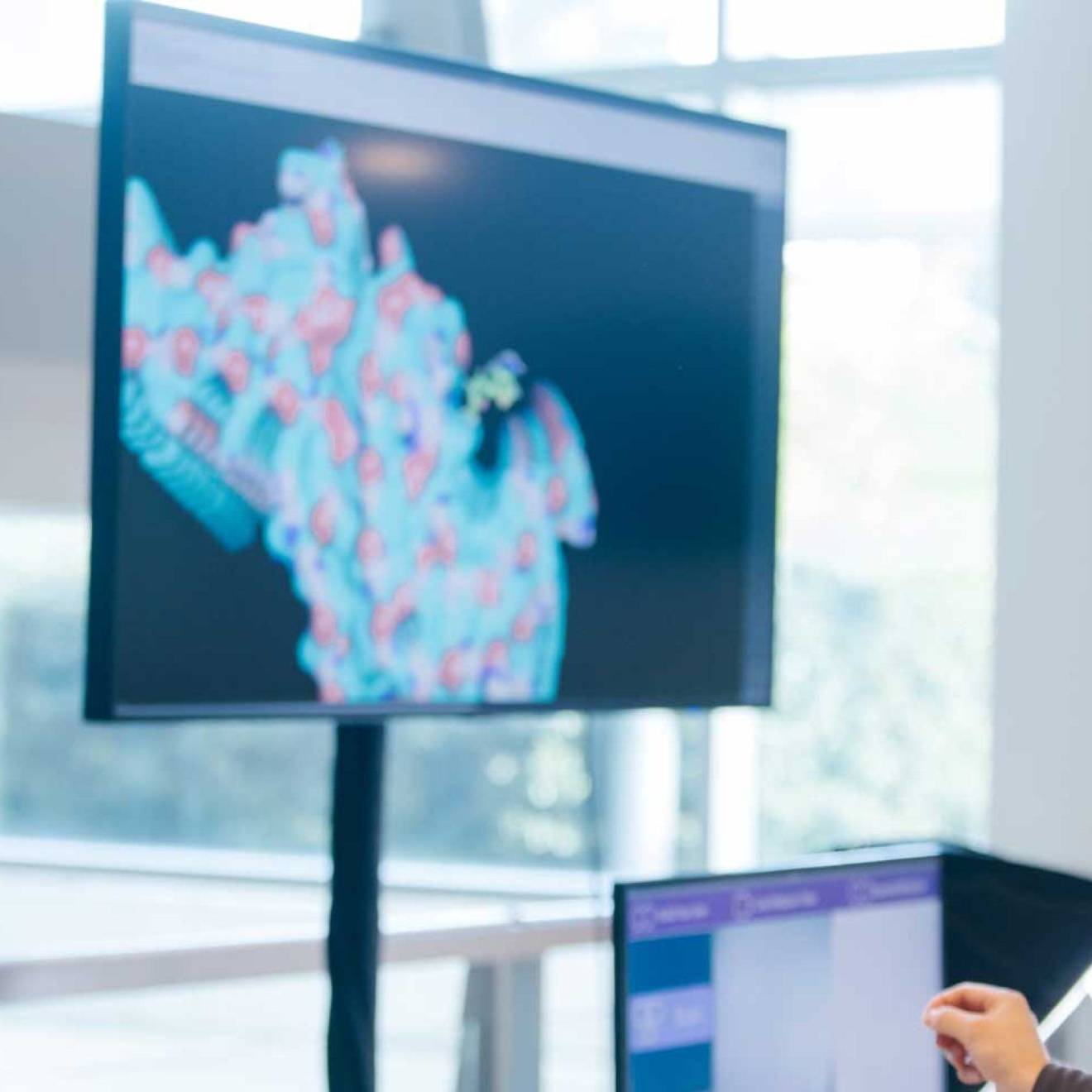 Someone wearing a virtual reality headset in front of two large screens showing molecules and one smaller screen