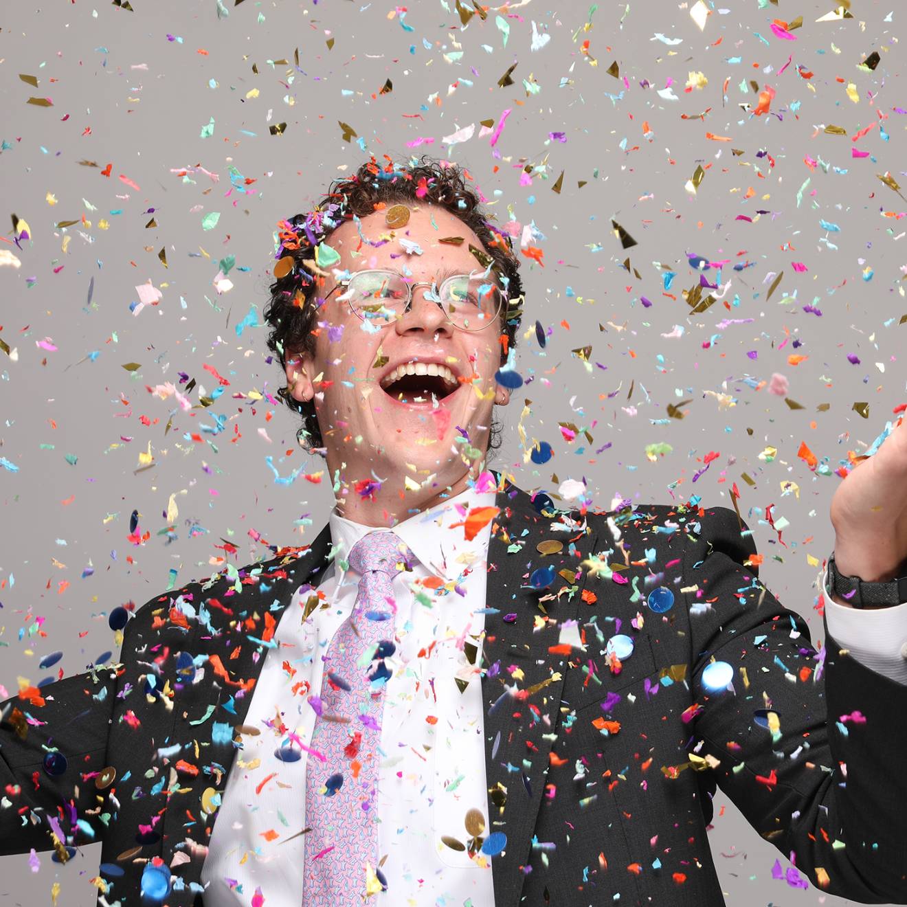 An ecstatically smiling man with glasses in a suit with a pink tie holds up his arms while confetti falls