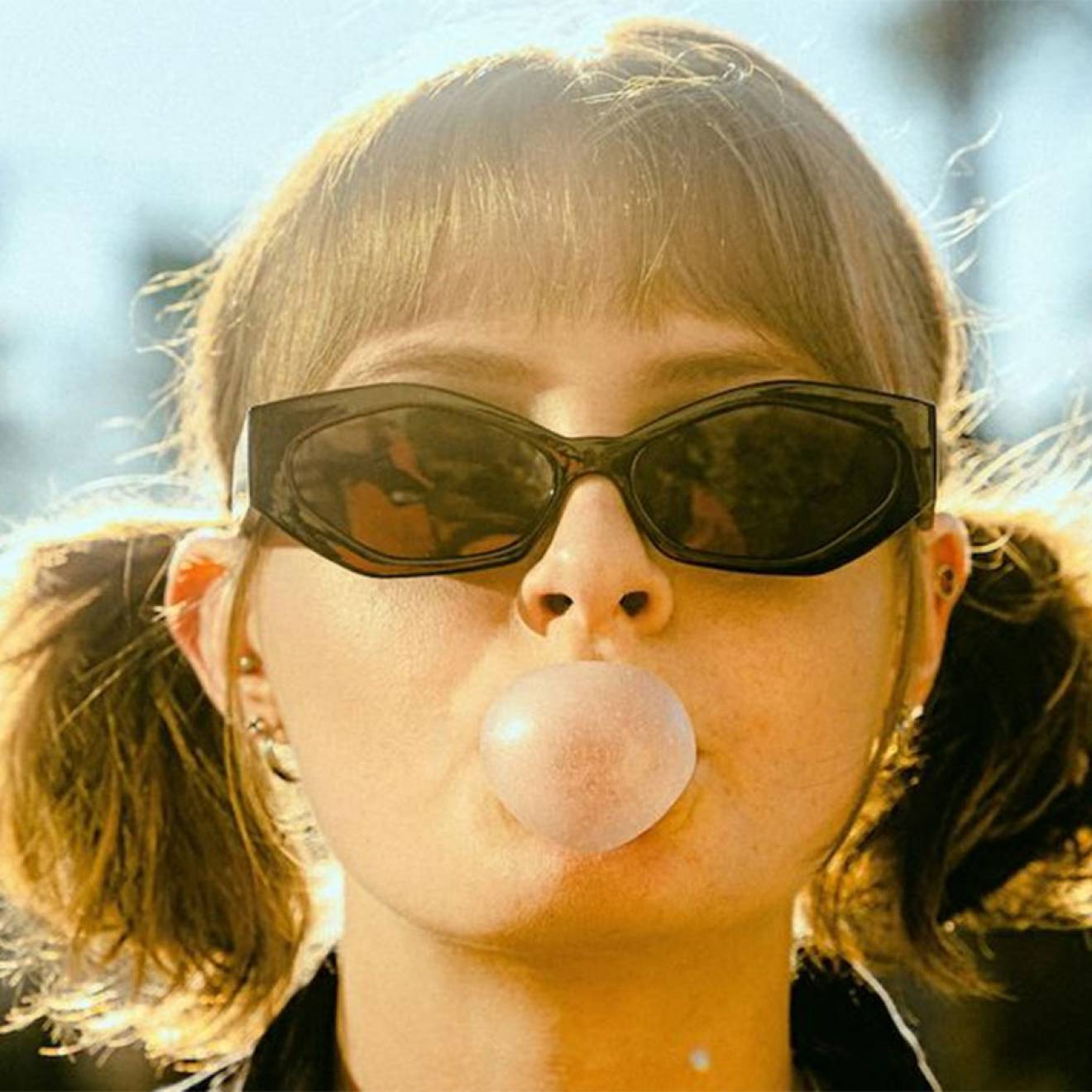 A woman wearing sunglasses and pigtails blows a gum bubble facing the camera