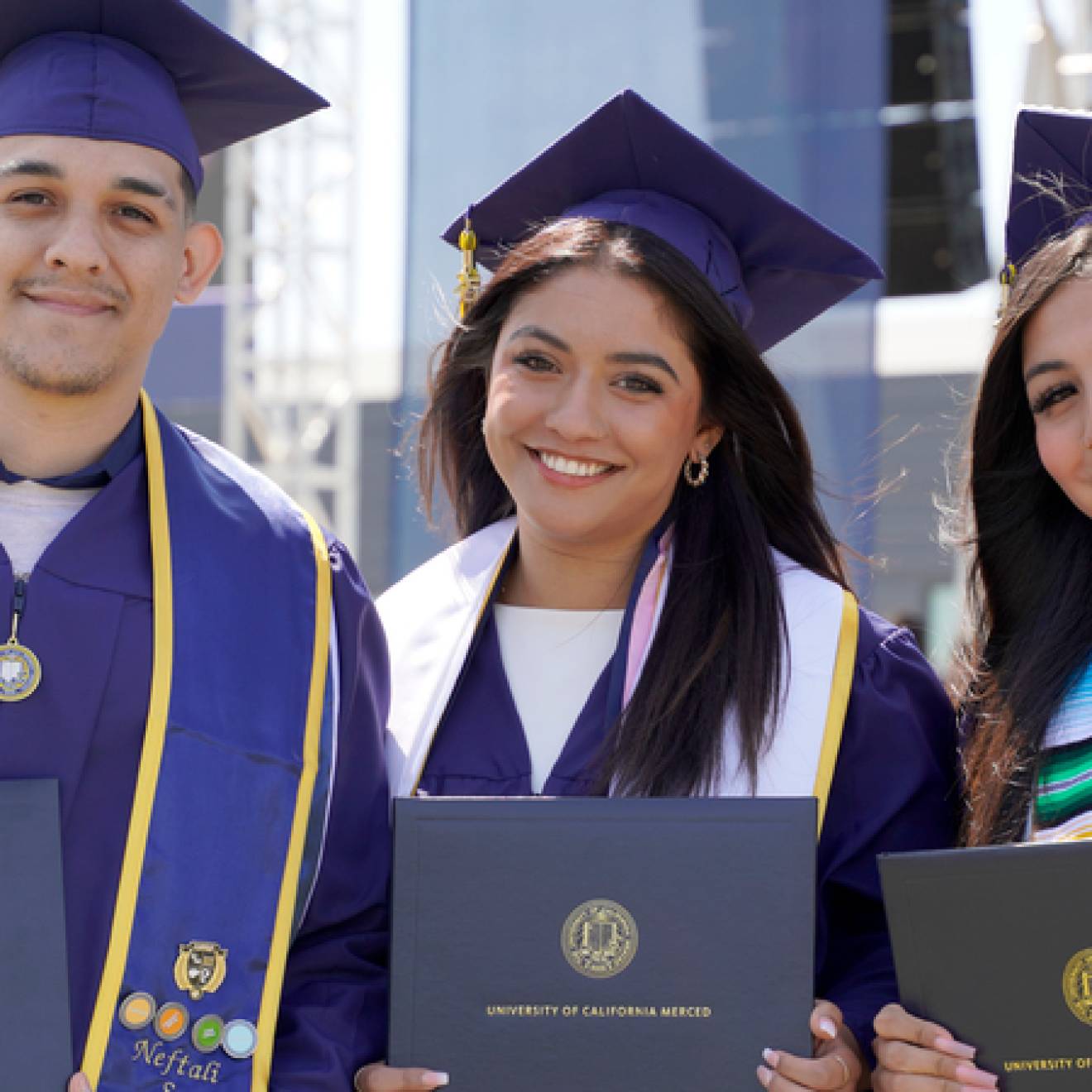 Three graduates in blue caps and gowns smile while holding their diplomas during a University of California, Merced commencement ceremony. They stand side by side, wearing stoles and tassels, with a stage and other graduates visible in the background.