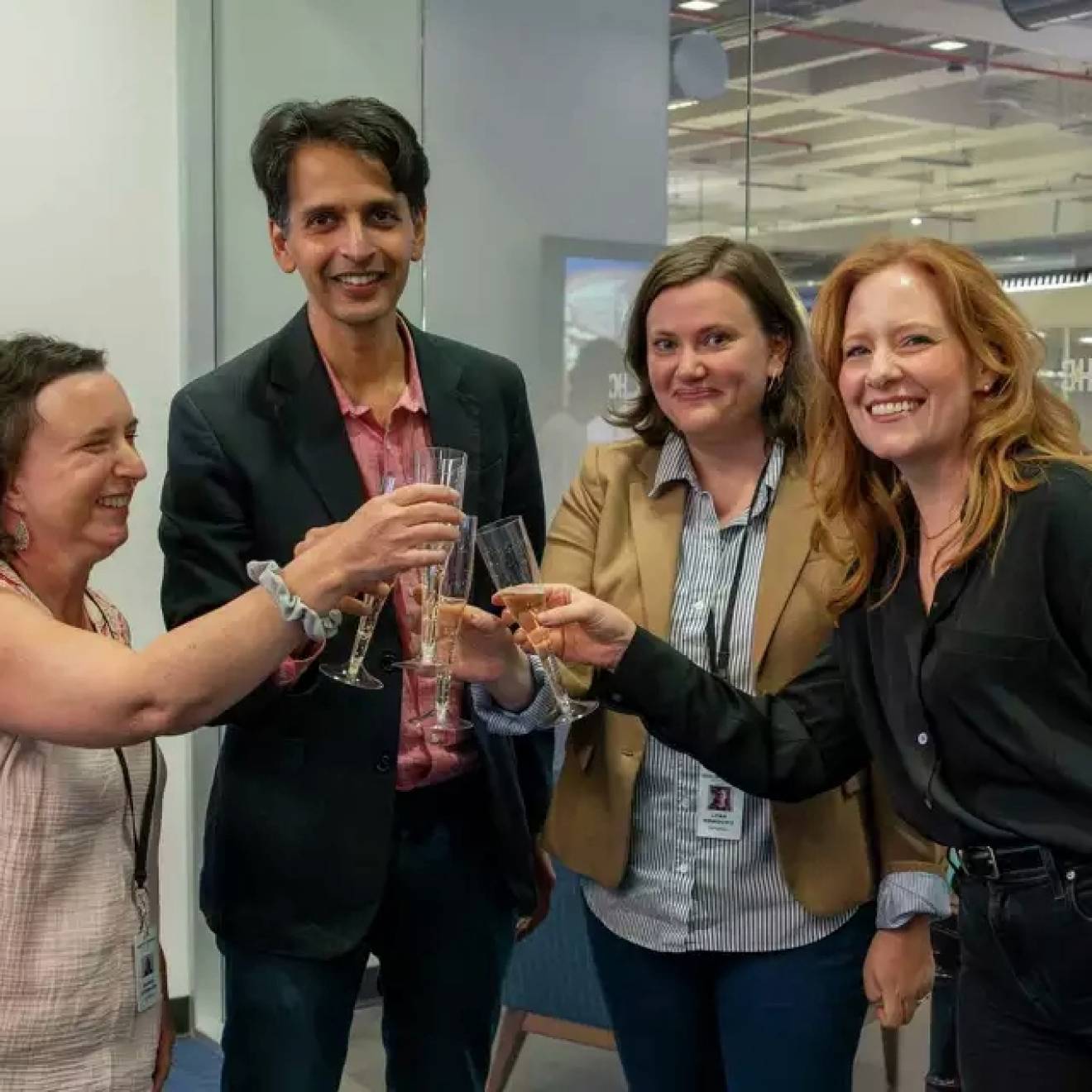 Four people in a newsroom do a champagne toast, smiling at the camera