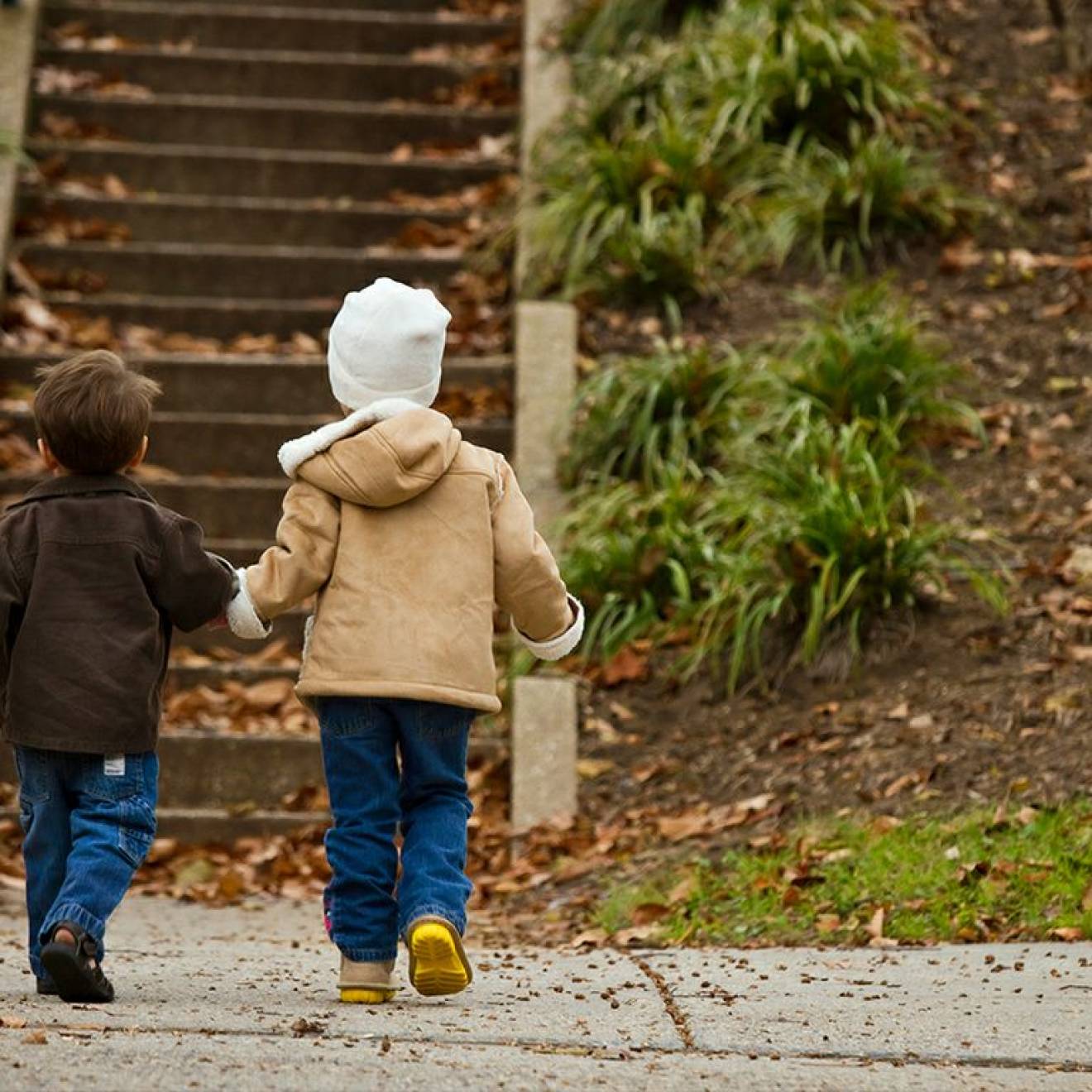 Two young children in winter clothes hold hands as they approach a park stairway, photographed from behind