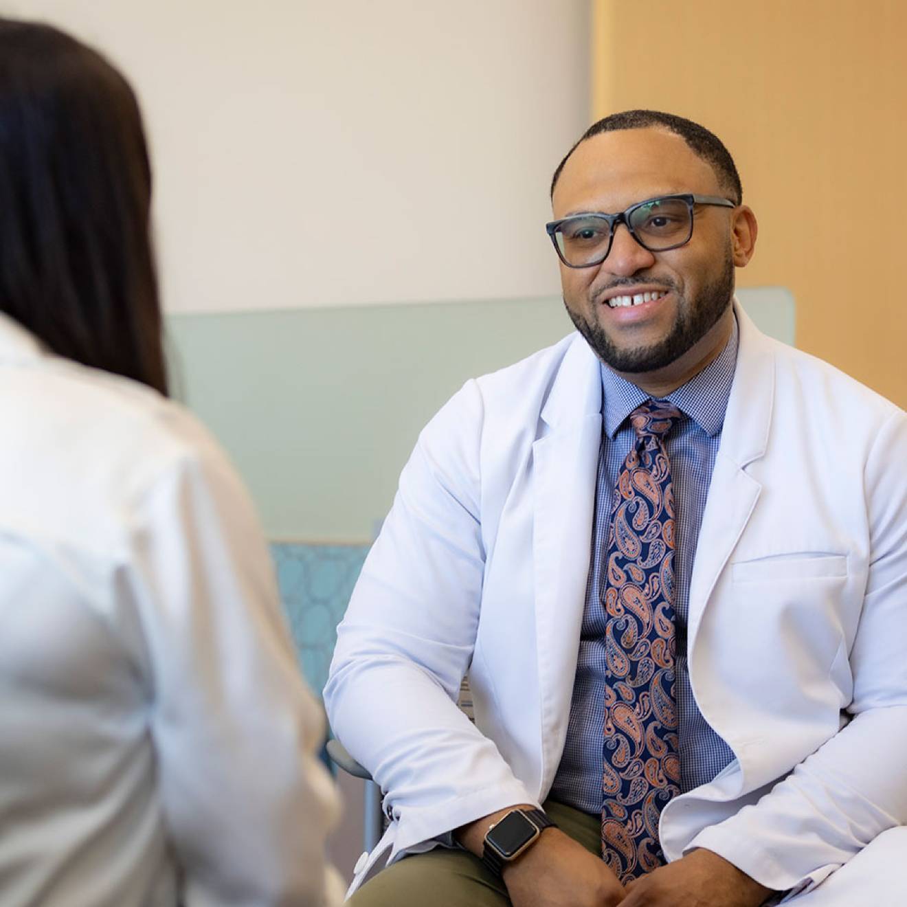 Chris Hall wearing a white medical provider's coat speaks with a patient in a lobby