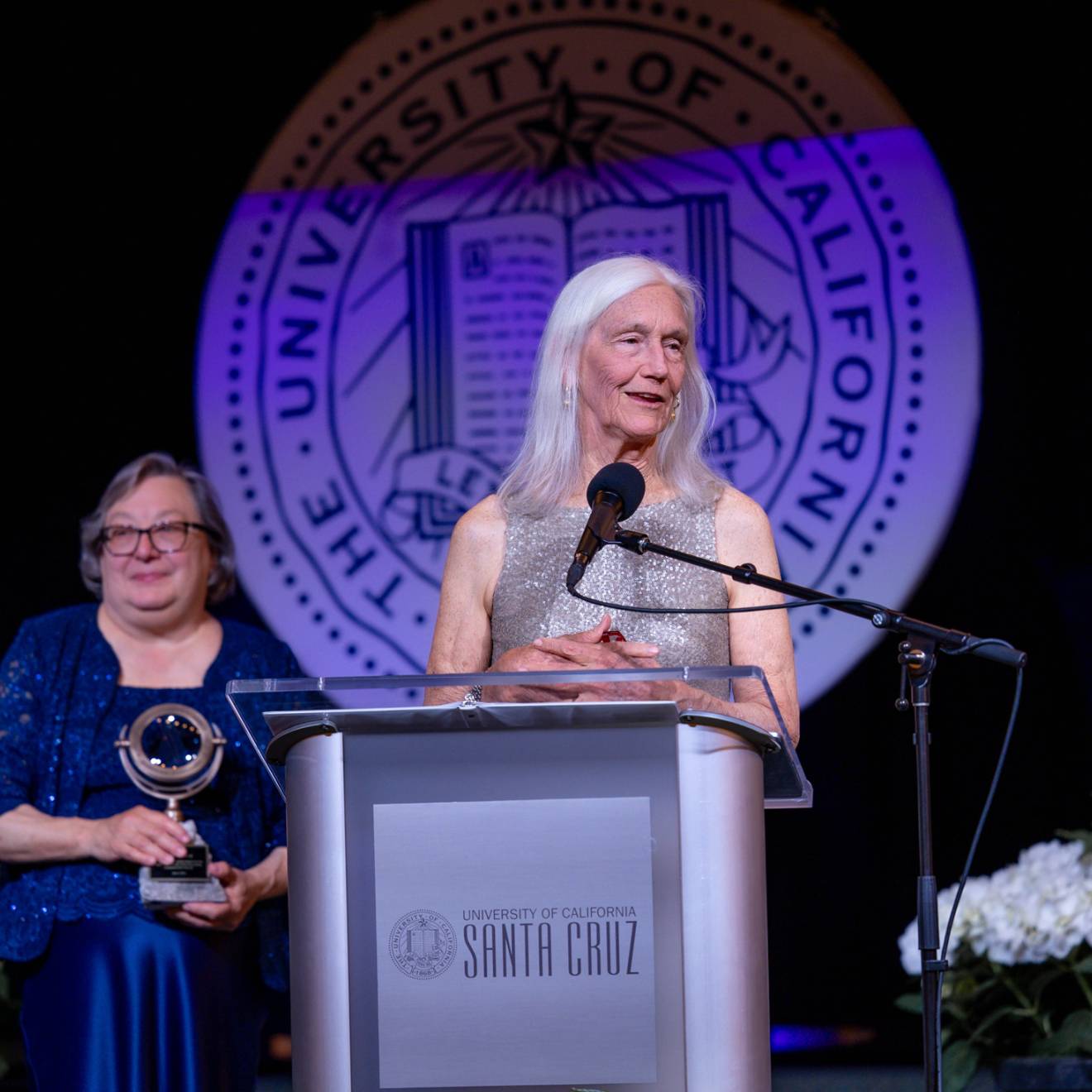 Woman with long white hair (Julie Packard) in silvery dress at a podium; woman in blue dress with short gray hair, Chancellor Larive, holding an award, stands behind