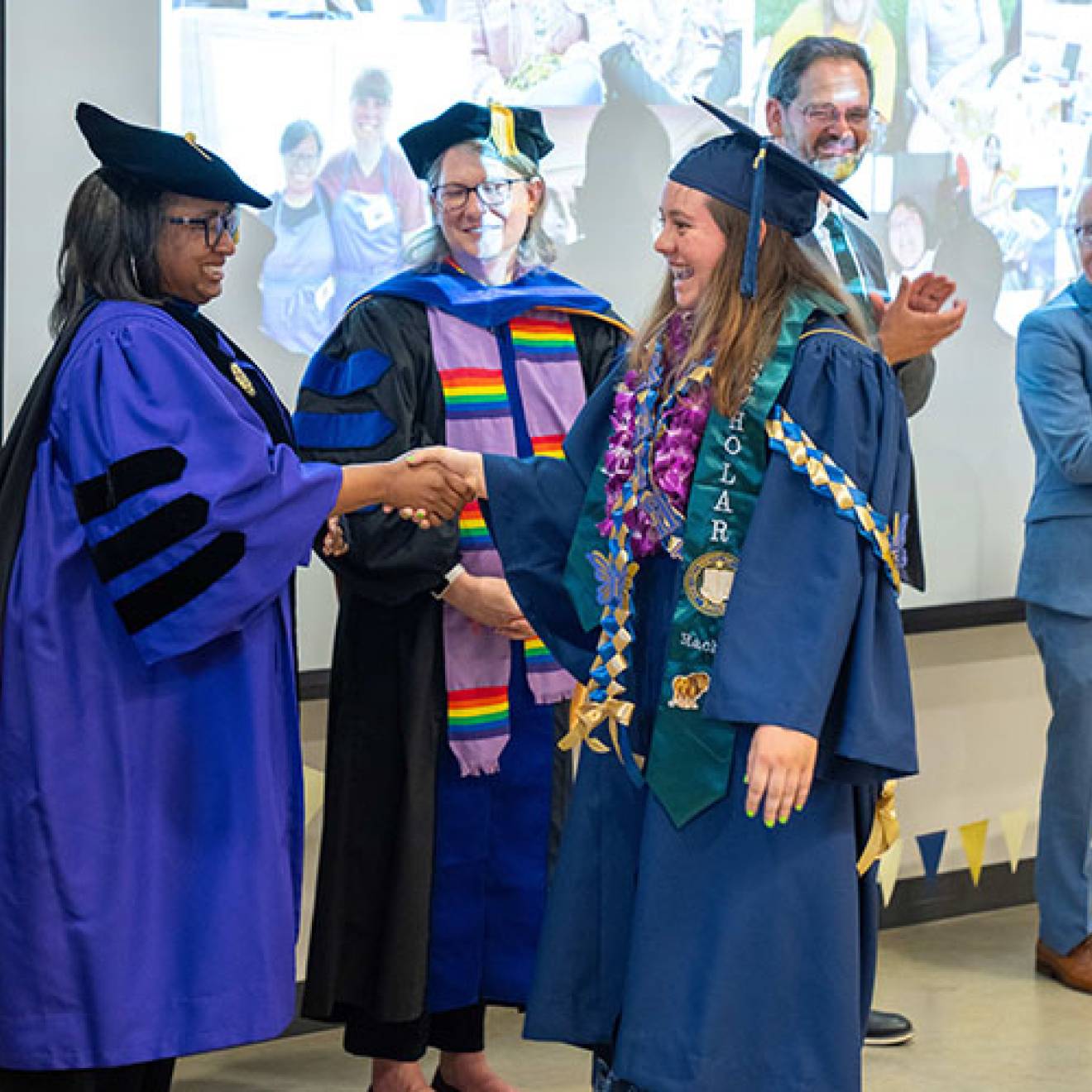A UC Davis graduate in graduation robe and cap receiving her diploma from university leaders dressed in academic regalia
