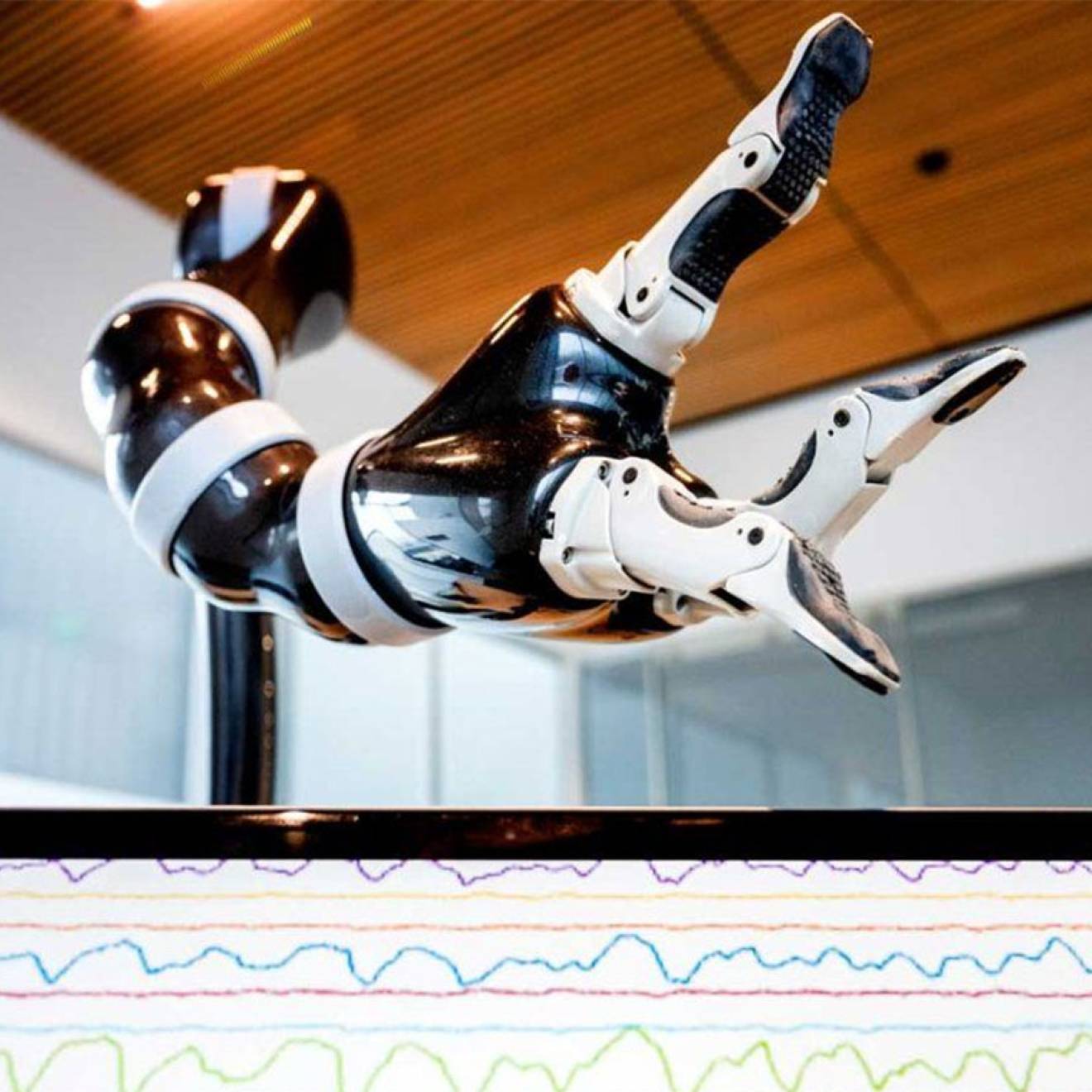 A robotic arm hovers over a computer screen showing brainwaves as a stack of tiny lines. 