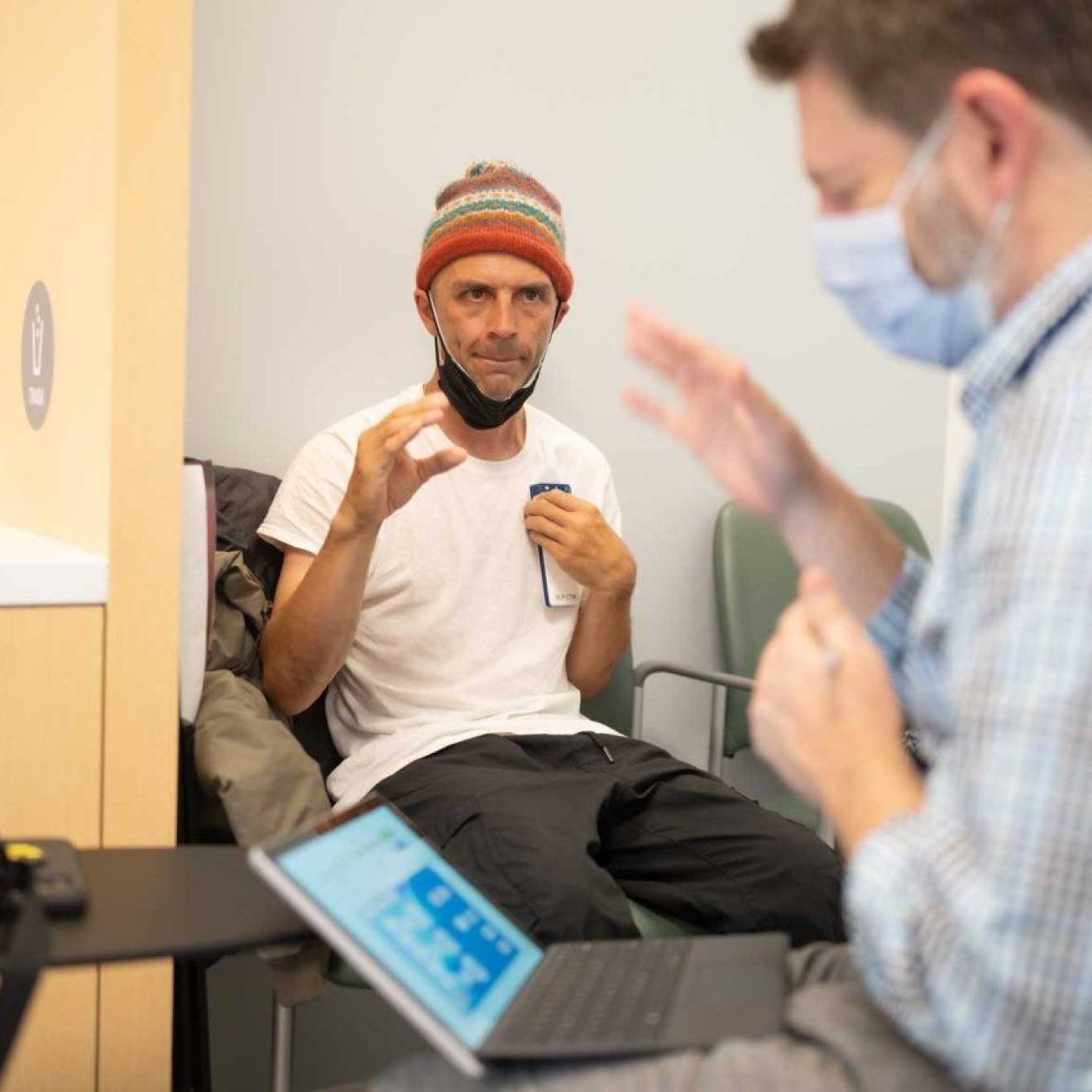 In the background, in focus, a man in a beanie makes a gesture with his hand as if to grasp. In the foreground, a doctor in a mask in front of a computer screen makes the same gesture