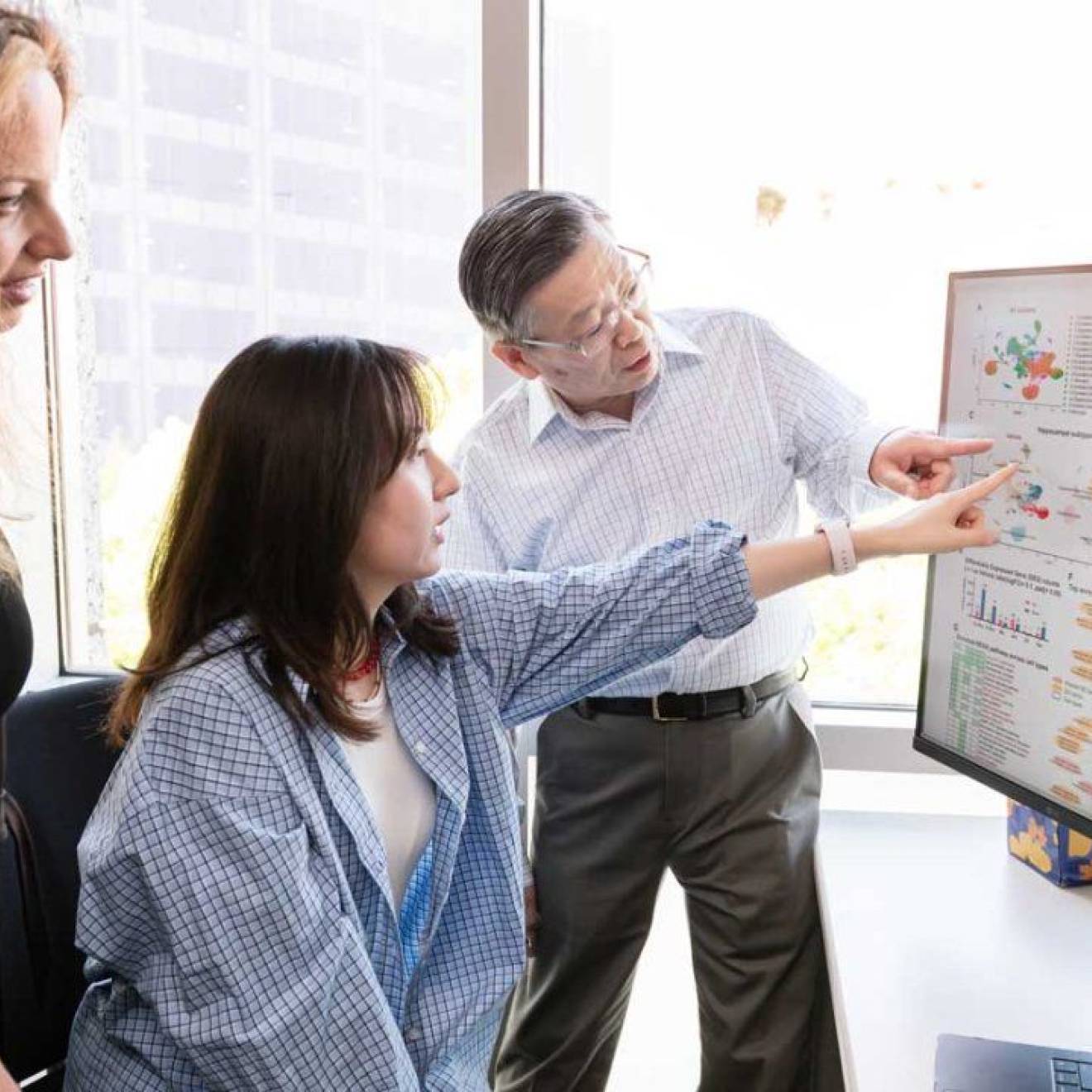 Three people in an office look at a large screen next to another large screen