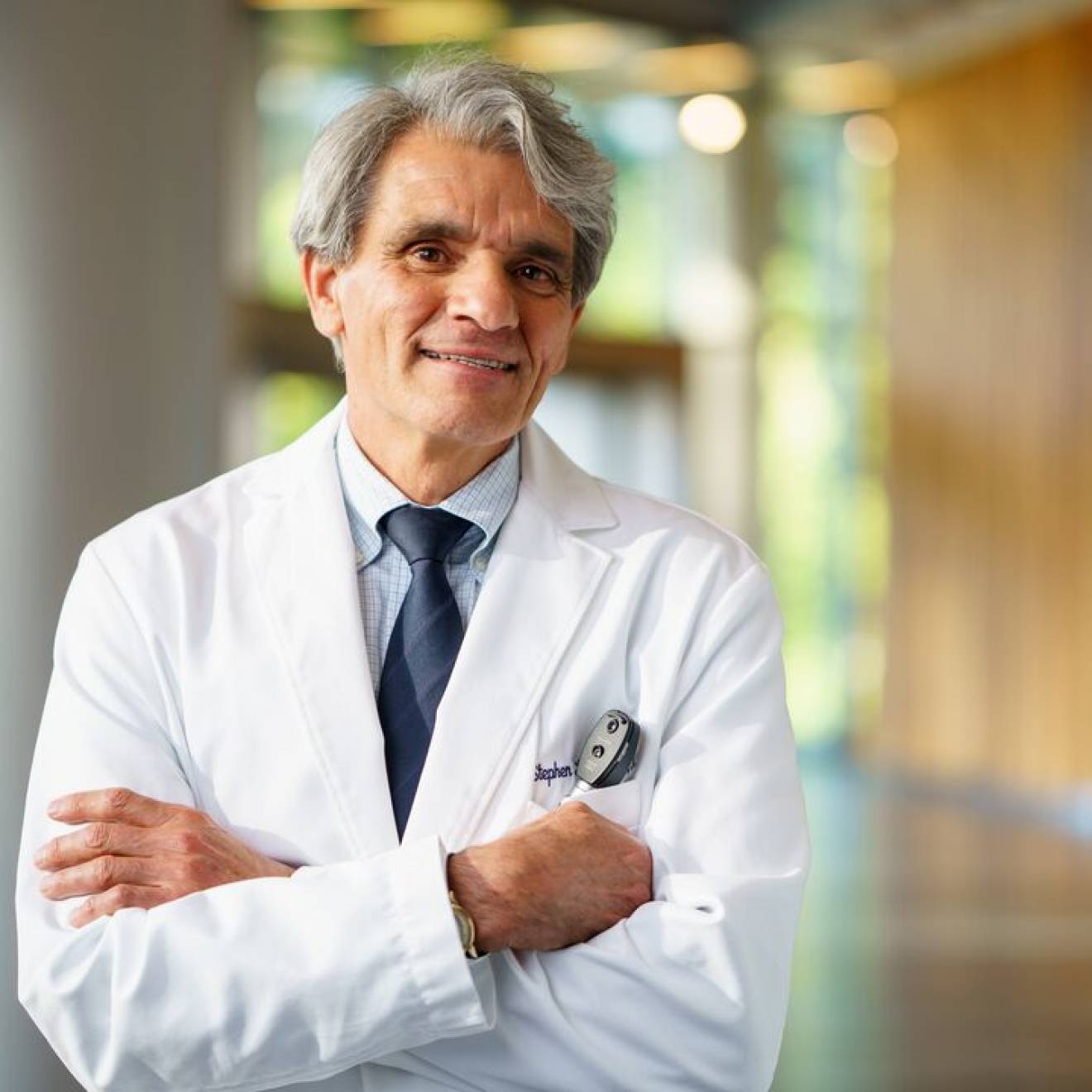 Man with white hair and arms crossed in a white doctor's coat smiles at camera in a hallway