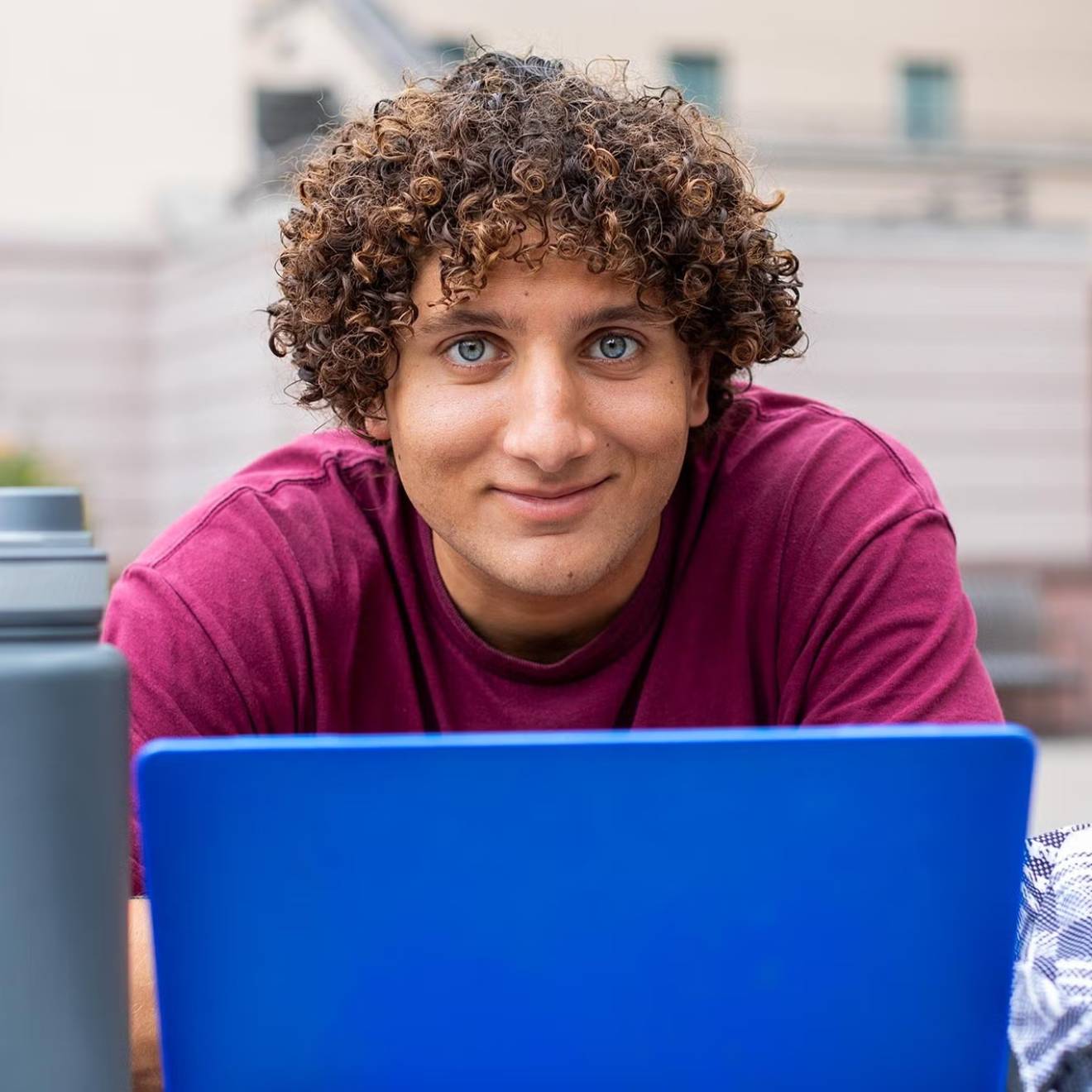 A young man with curly hair stares intently above a laptop while sitting outdoors