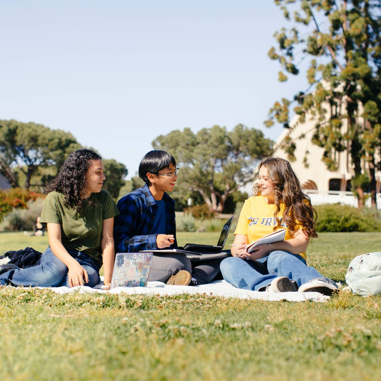 Three students on a picnic blanket studying talk to each other on the UC Irvine campus under a bright sky