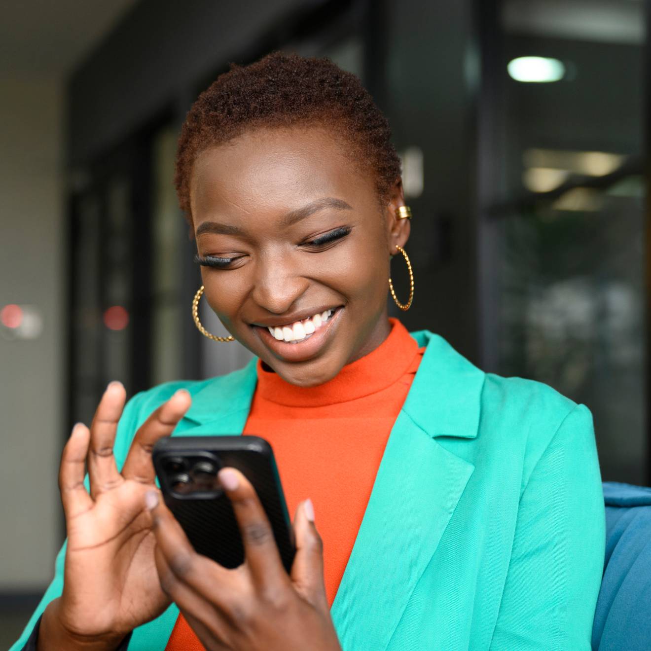 Waist-up view of shorthaired Black woman wearing hoop earrings, teal jacket over orange top, and smiling while scrolling portable device in modern office